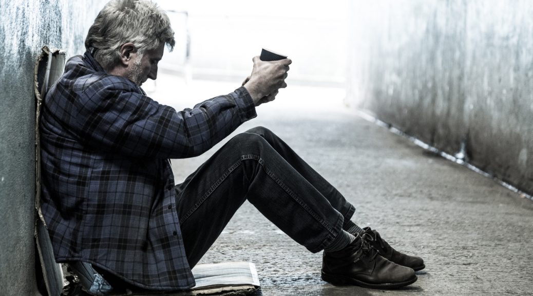 Homeless man outside sitting against wall with cup of coffee
