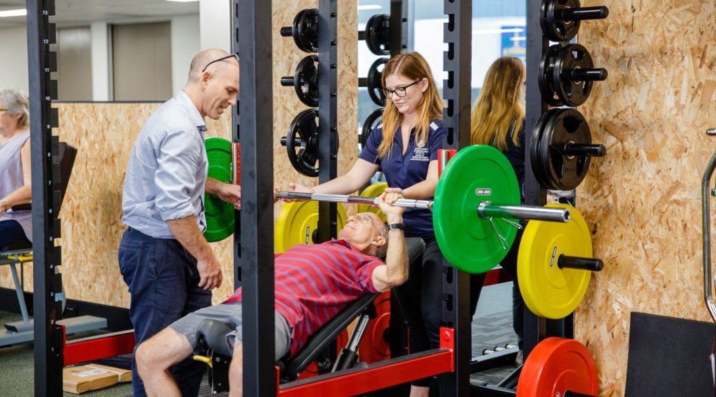 UQ Healthy Living gym, client on bench press machine being assisted by physiotherapist and student.